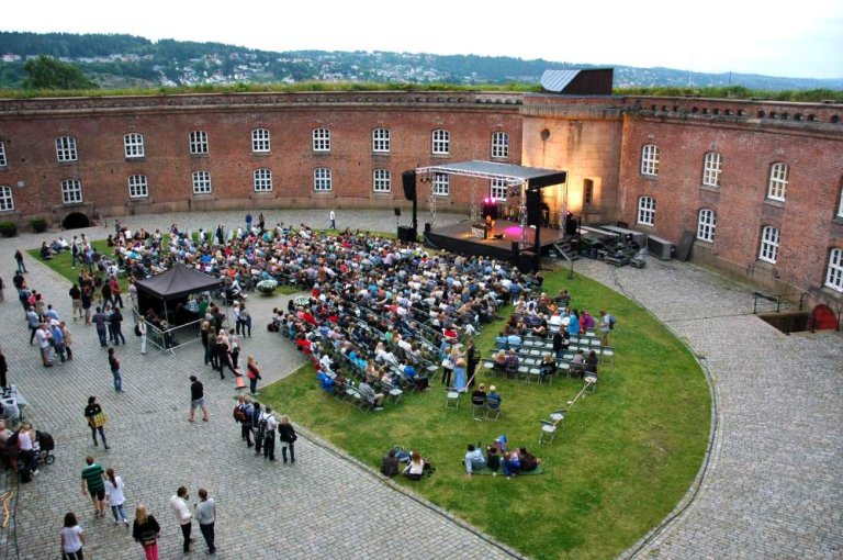 Utendørs arrangement i borggården med scene, lys- og lydutstyr, og et stort publikum foran scenen.