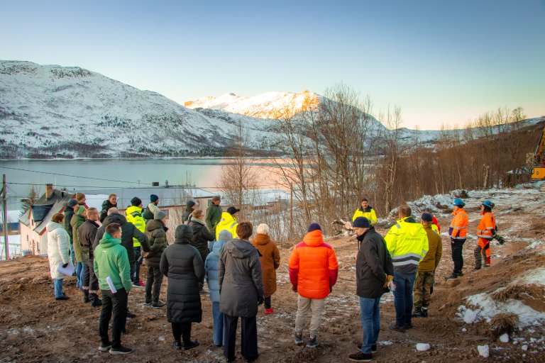 Gruppe med mennesker som er samlet for markering av første spadetak på ny befalsforlenging. Utsikt over fjorden. Snø, kaldt sol. Trær og brunt gress. 