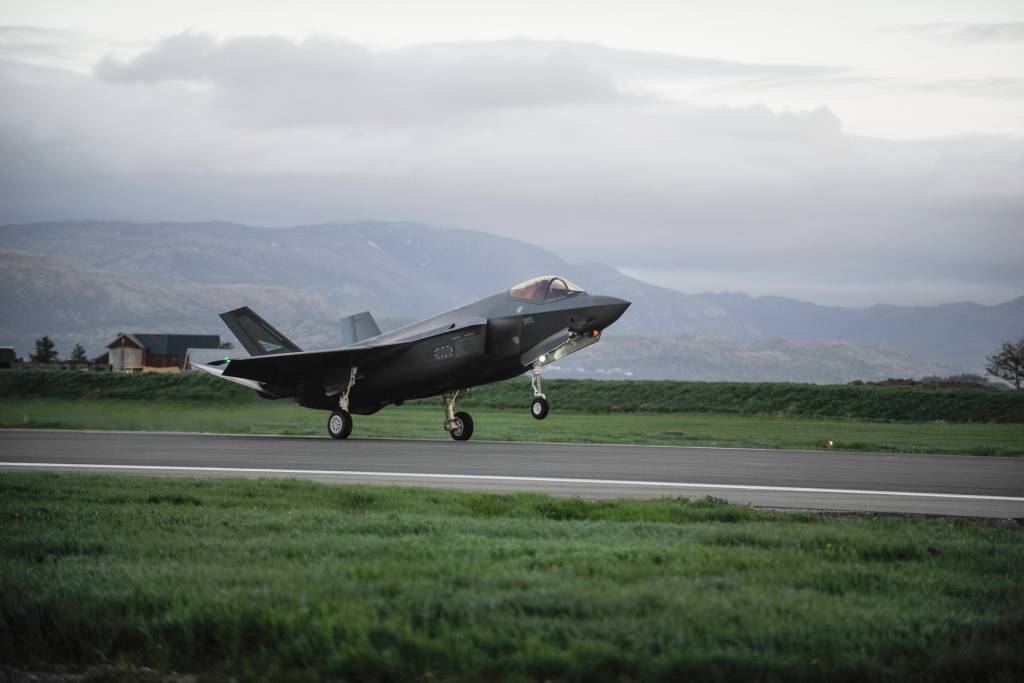 ET F-35 kampfly er i ferd med å ta av fra en flystripe omkranset av grønt gress. I bakgrunnen er et gårdsbruk og fjell. Himmelen er grå og skyet.